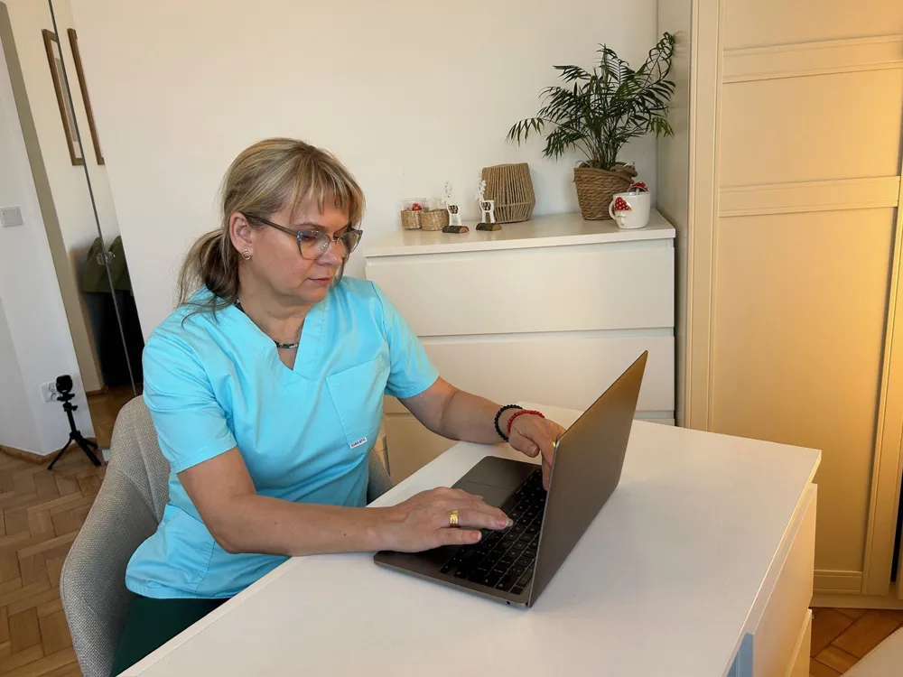 Terapeutka w błękitnym stroju medycznym pracująca przy laptopie w jasnym, nowocześnie urządzonym gabinecie.